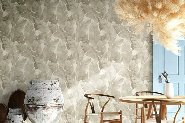Marburg Avalon 31625 Dining area with neutral leaf-pattern wallpaper, a round wooden table and chairs, a blue door, a large rustic ceramic urn, a feathery pendant light, and a small vase with branches.