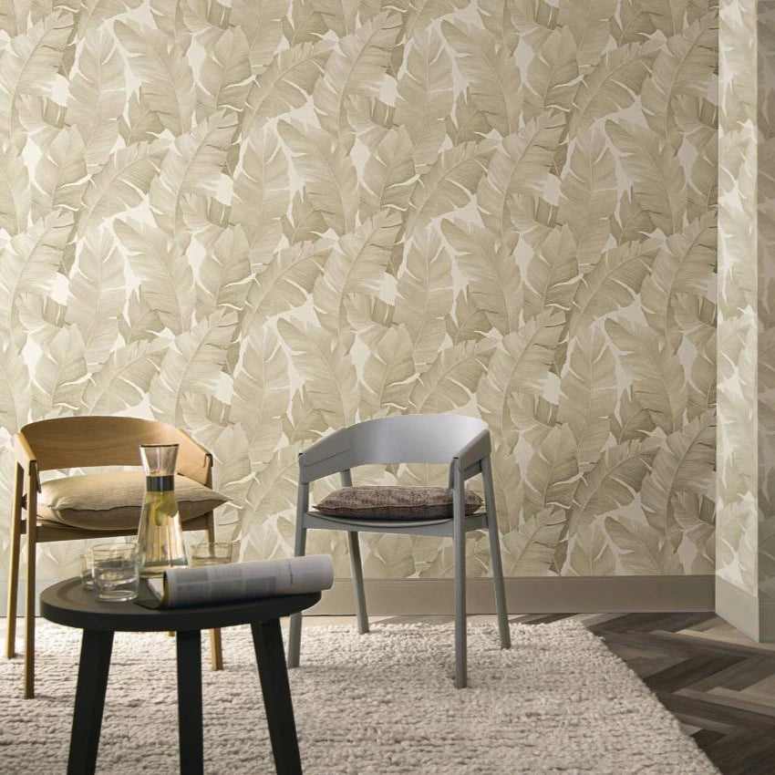 Marburg Avalon 31625 Beige tropical leaf wallpaper on two walls behind a seating corner with two modern chairs, a small round table holding a carafe, glasses, and a book, over a textured rug.