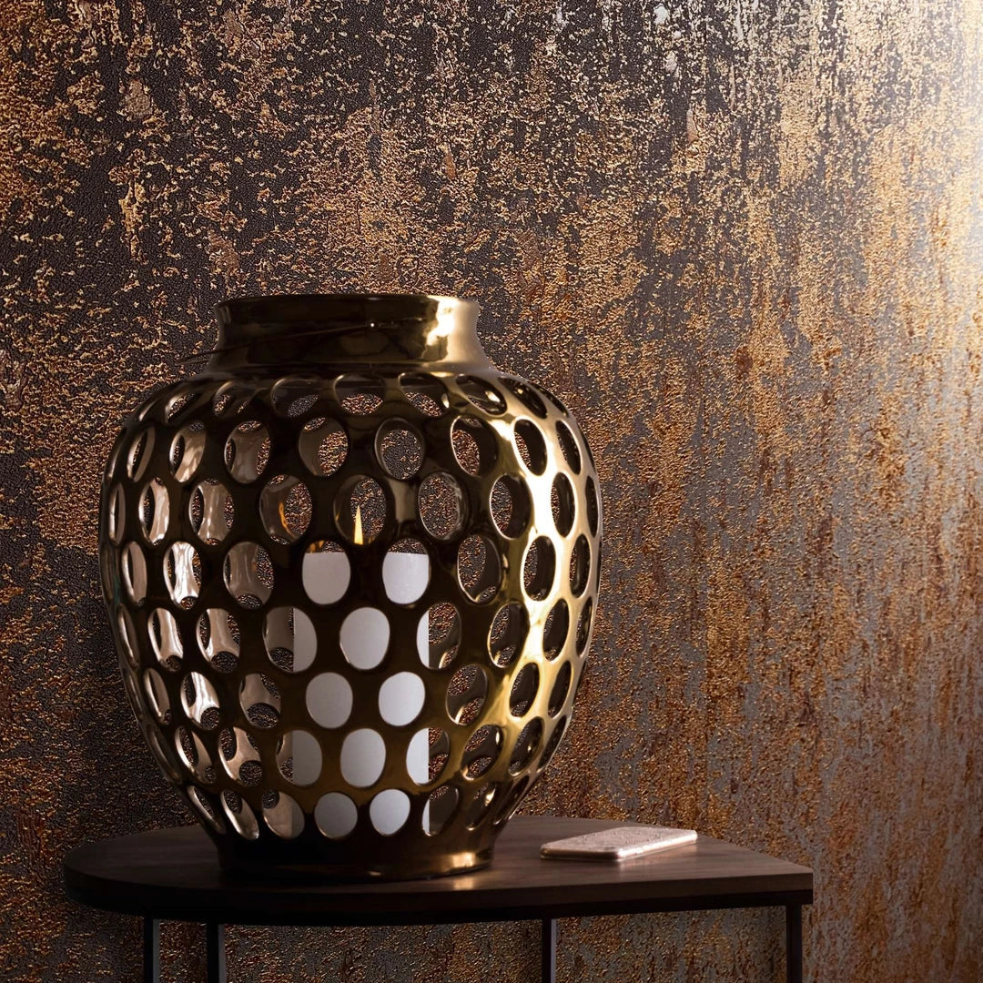 Marburg Avalon 31644 Close-up of copper and dark abstract textured wallpaper with irregular vertical mottling, shown behind a perforated metal lantern holding a candle on a small side table with a rectangular item.