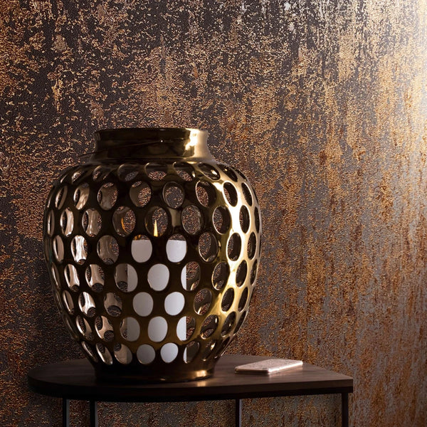 Marburg Avalon 31644 Close-up of copper and dark abstract textured wallpaper with irregular vertical mottling, shown behind a perforated metal lantern holding a candle on a small side table with a rectangular item.