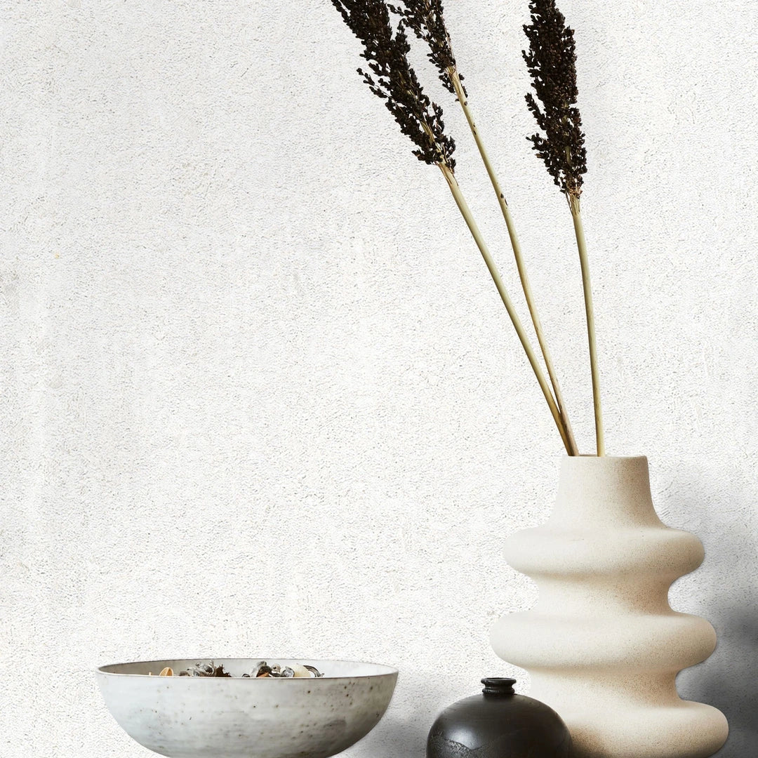 Marburg Avalon 31645 White textured wallpaper with subtle mottling behind a wavy sculptural ceramic vase with tall dried stems, a small dark vase, and a shallow bowl.