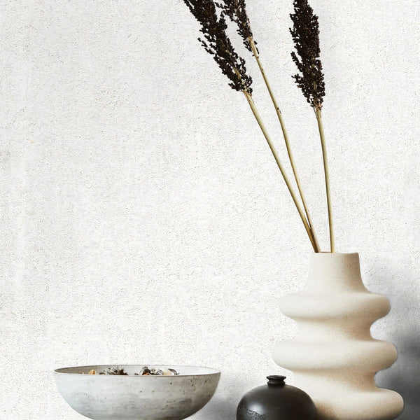 Marburg Avalon 31645 White textured wallpaper with subtle mottling behind a wavy sculptural ceramic vase with tall dried stems, a small dark vase, and a shallow bowl.