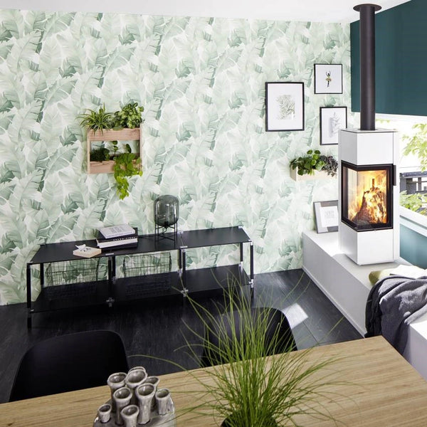 Marburg Avalon 31650 Dining area featuring a soft green and white leaf-pattern wallpaper, a modern white wood‑burning stove, a black console table with framed art above, and several plants, with a wooden table and black chairs in the foreground.