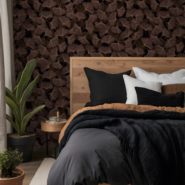 Marburg Boho 35779 Bedroom scene with a dark leaf-pattern wallpaper of overlapping brown fan-shaped leaves behind a wood headboard, with a bed, round side table, and potted plants in view.