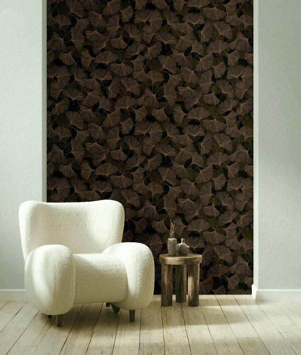 Marburg Boho 35779 Dark brown ginkgo leaf wallpaper on a feature wall behind a white armchair and a small wooden side table on light wood flooring.
