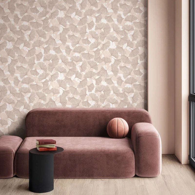 Marburg Boho 35780 Beige and light brown ginkgo leaf wallpaper behind a modern curved sofa, with a small black side table holding books, a round pillow on the sofa, and a window beside the scene.