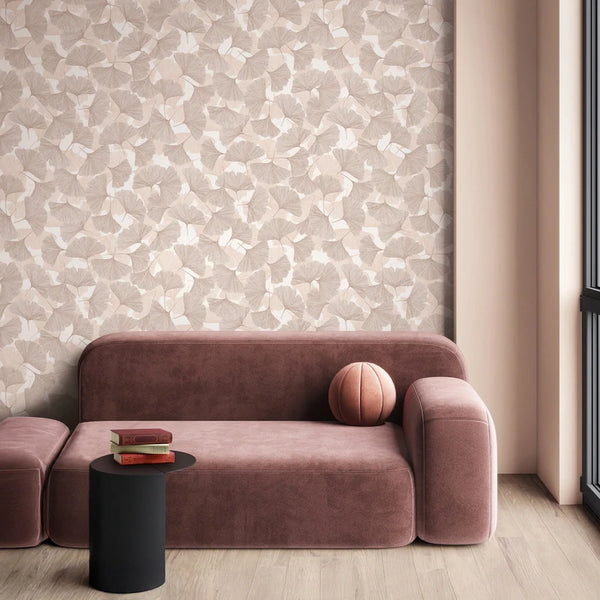 Marburg Boho 35780 Beige and light brown ginkgo leaf wallpaper behind a modern curved sofa, with a small black side table holding books, a round pillow on the sofa, and a window beside the scene.