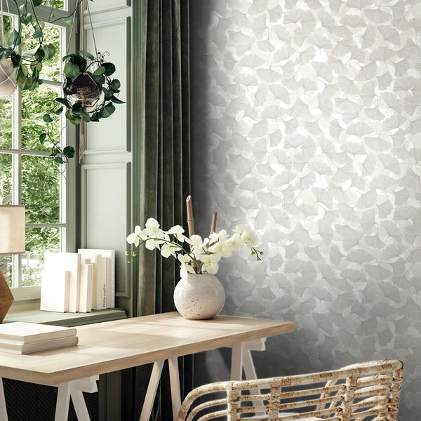 Marburg Boho 35781 Grey and white leaf-patterned wallpaper on a wall beside a window with green curtains, styled with a light wood desk, chair, books, and a vase of white flowers.