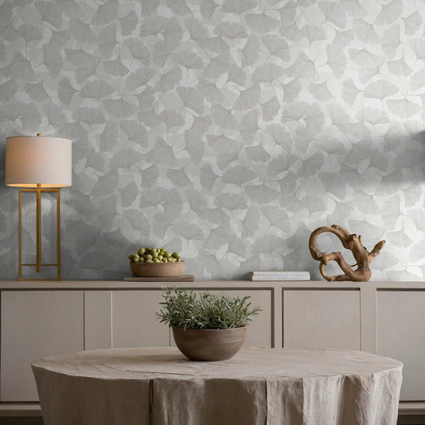 Marburg Boho 35781 Grey and white ginkgo leaf wallpaper behind a sideboard holding a table lamp, books, a decorative wooden sculpture, and a bowl of green fruit, with a round draped table and a potted plant in the foreground.