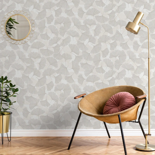 Marburg Boho 35782 Beige and grey ginkgo leaf wallpaper shown in a styled room with a tan lounge chair holding a round pink cushion, a gold floor lamp, a small round mirror, and a potted plant on a stand.