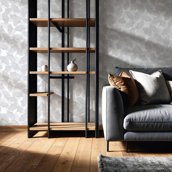 Marburg Boho 35783 Gray leaf-pattern wallpaper shown in a living room scene behind a black metal and wood shelving unit and a gray sofa with pillows, on a wood floor with a rug.