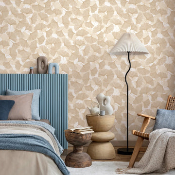 Marburg Boho 35784 Beige ginkgo leaf wallpaper behind a styled bedroom with a blue ribbed headboard, pillows and blankets, a curving floor lamp with a pleated shade, small side tables with ceramic decor, a wooden stool with an open book, and a woven chair.
