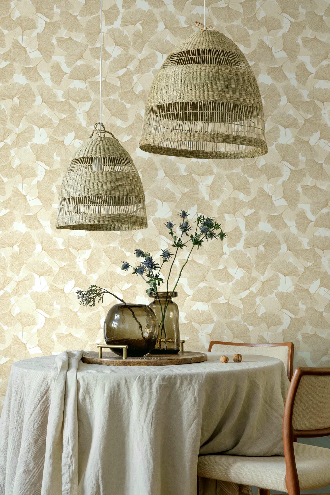 Marburg Boho 35784 Beige ginkgo leaf wallpaper behind a dining setup with two woven pendant lights, a round table draped in linen, glass vases with stems, and wood-framed chairs.