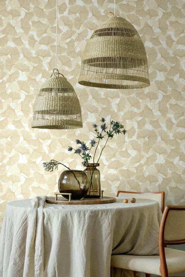 Marburg Boho 35784 Beige ginkgo leaf wallpaper behind a dining setup with two woven pendant lights, a round table draped in linen, glass vases with stems, and wood-framed chairs.