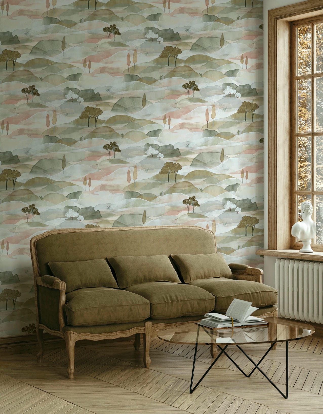 Marburg Boho 35785 Scenic watercolor hills wallpaper with small stylized trees in muted green and blush tones, shown behind a cushioned sofa, a glass coffee table with an open book, and a window with a white sculptural bust on the sill.