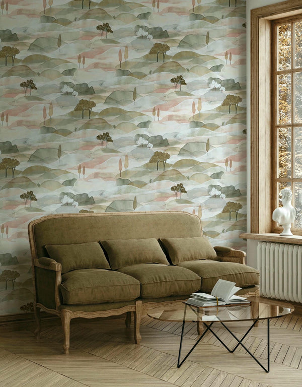 Marburg Boho 35785 Scenic watercolor hills wallpaper with small stylized trees in muted green and blush tones, shown behind a cushioned sofa, a glass coffee table with an open book, and a window with a white sculptural bust on the sill.