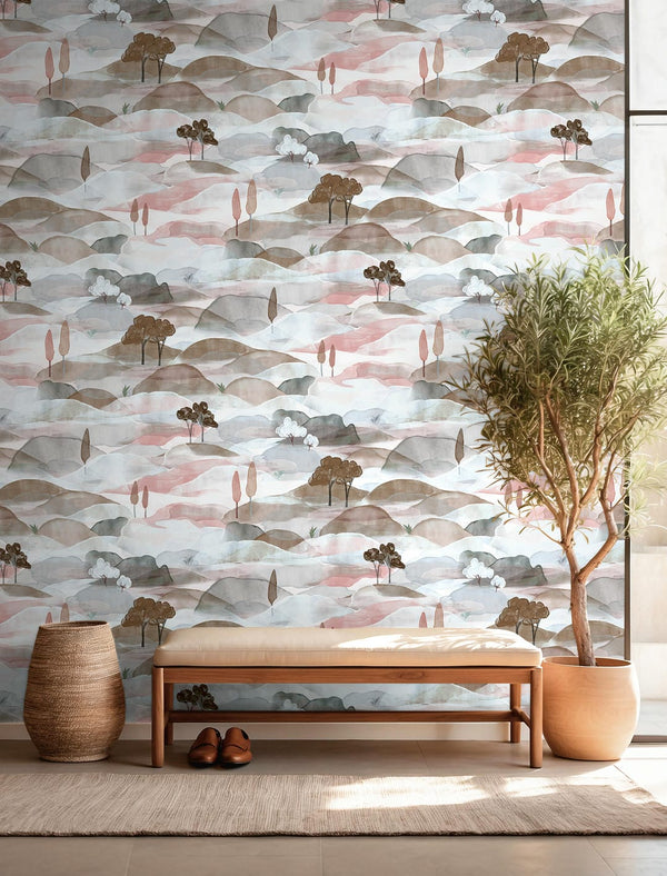 Marburg Boho 35786 Watercolor landscape wallpaper with layered hills and small trees in muted pink, taupe, and blue-gray, shown with a wooden bench, woven basket, rug, shoes, and a potted tree by a window.