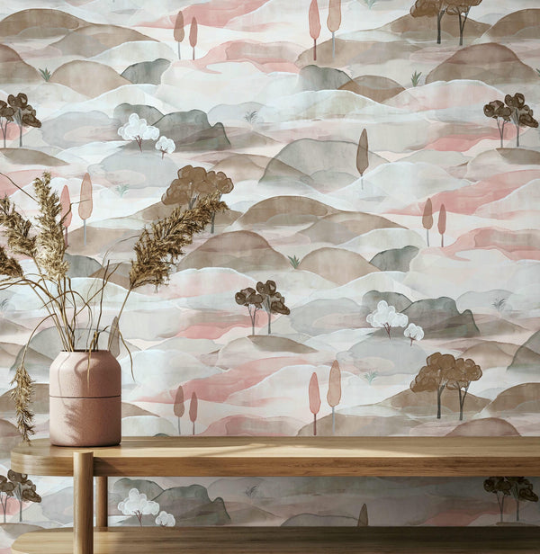 Marburg Boho 35786 Watercolor hills and trees wallpaper in soft pink, taupe and muted green, with a wooden console and a pink ceramic vase holding dried grasses in front.