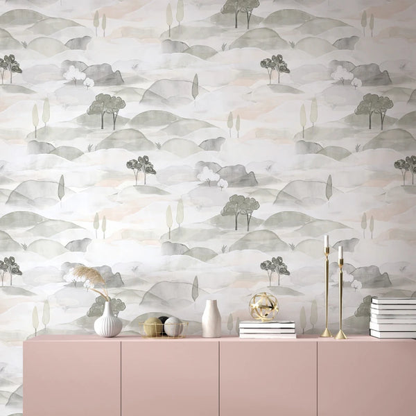 Marburg Boho 35787 Watercolor-style landscape wallpaper with soft grey and green hills and small trees, shown behind a pink sideboard styled with vases, dried grass, gold candlesticks, stacked books, and a gold geometric orb.