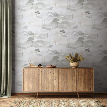 Marburg Boho 35787 Grey and white watercolor landscape wallpaper with layered hills and simple trees behind a wood sideboard. A vase with dried grasses, a bowl, a small jar, a wooden tray, a green curtain, and a textured rug are visible.