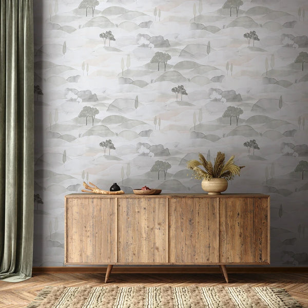 Marburg Boho 35787 Grey and white watercolor landscape wallpaper with layered hills and simple trees behind a wood sideboard. A vase with dried grasses, a bowl, a small jar, a wooden tray, a green curtain, and a textured rug are visible.