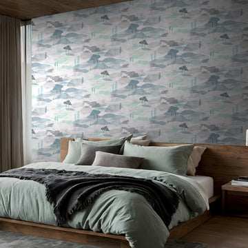 Marburg Boho 35788 Watercolor landscape wallpaper with layered hills and small trees in soft blue, green, and gray on the wall behind a bed in a modern bedroom.