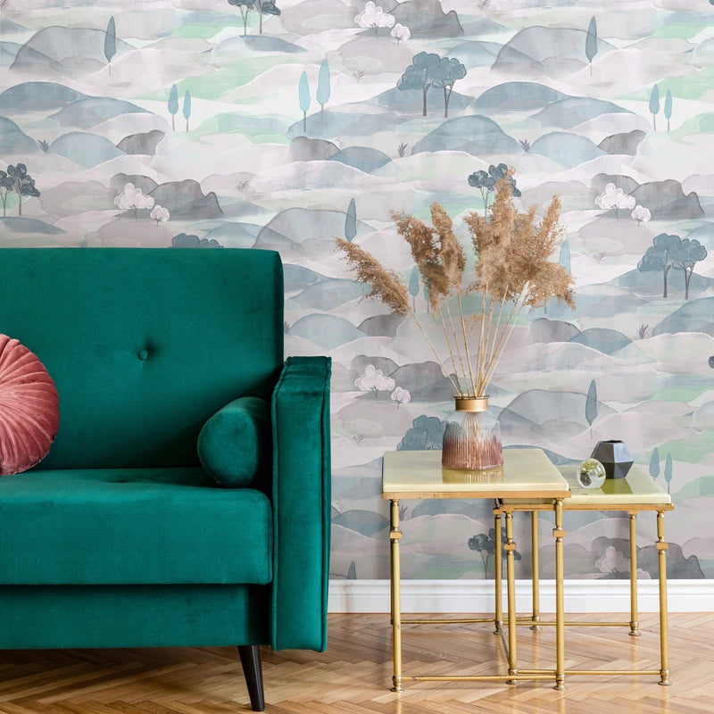 Marburg Boho 35788 Landscape wallpaper with watercolor hills and small trees in blue, green, and grey behind a teal sofa and gold nesting tables holding a vase of dried grasses on a herringbone wood floor.