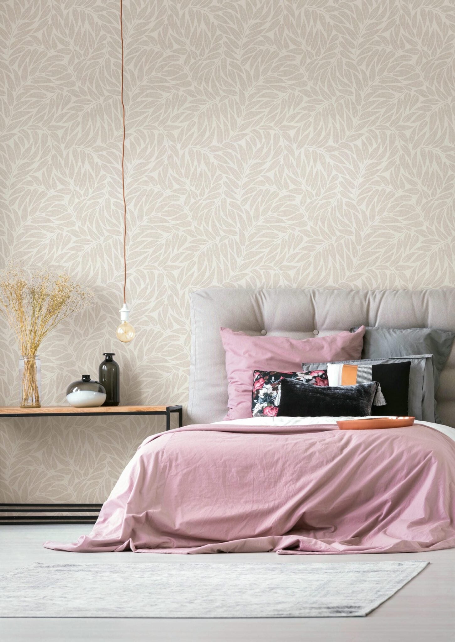 Marburg Boho 35792 Leaf-pattern wallpaper in light neutral tones shown behind a bed with pink bedding and a padded headboard, a side table with vases and dried stems, and a hanging bulb.