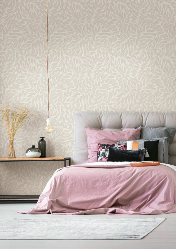 Marburg Boho 35792 Leaf-pattern wallpaper in light neutral tones shown behind a bed with pink bedding and a padded headboard, a side table with vases and dried stems, and a hanging bulb.