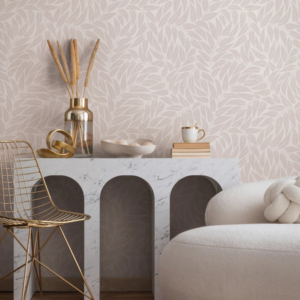 Marburg Boho 35792 Neutral leaf-pattern wallpaper behind a marble console with arched openings, a gold wire chair, a glass vase with dried stems, a small bowl, stacked books with a mug, and a cream upholstered armchair with a knot cushion.