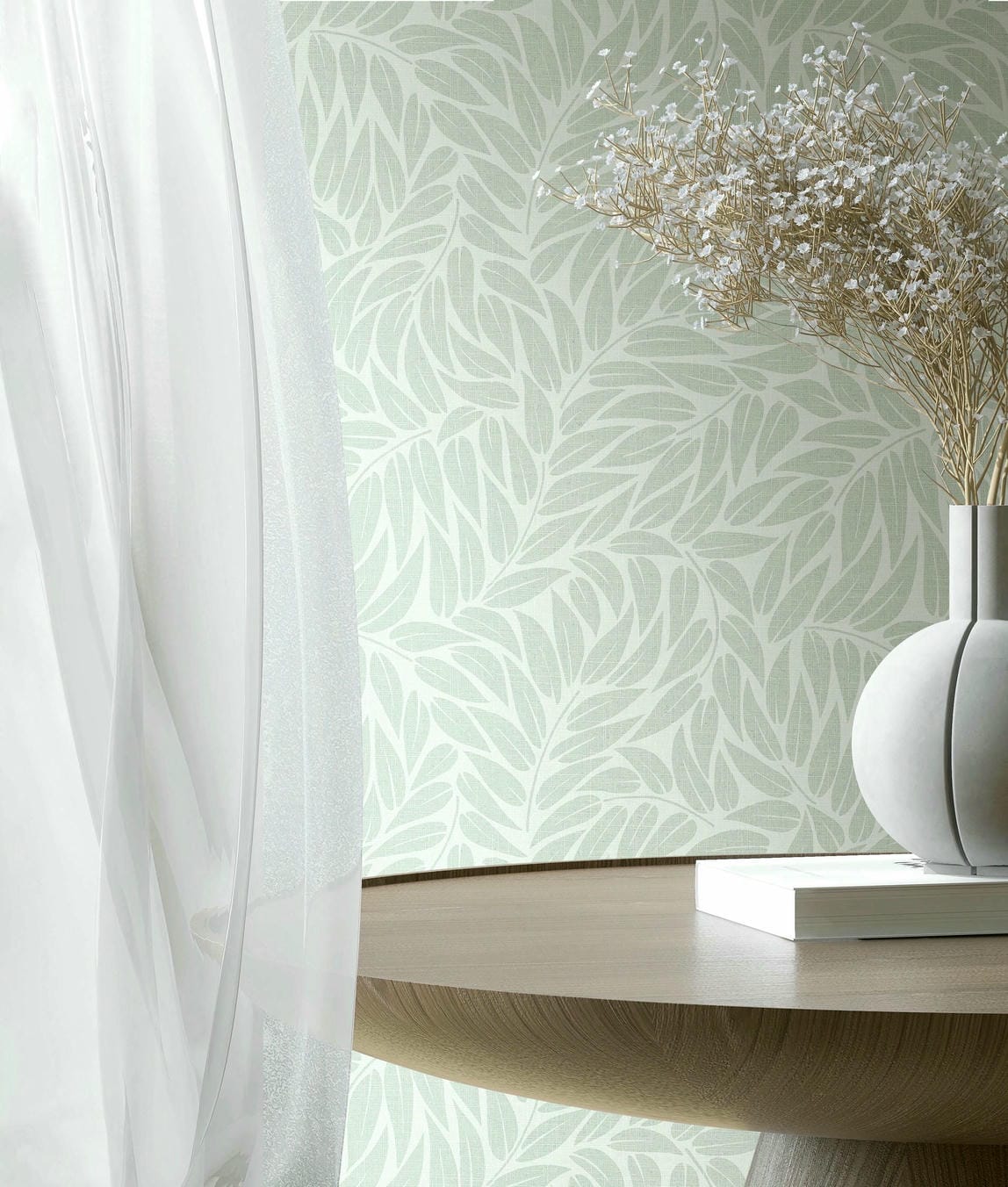 Marburg Boho 35793 Close-up of pale green leaf-pattern wallpaper behind a round wooden table with a white sculptural vase holding dried stems, a book, and a sheer curtain.