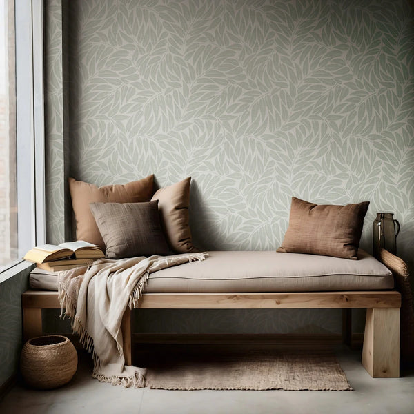 Marburg Boho 35793 Green leafy botanical wallpaper behind a wooden bench with neutral pillows and a throw by a window, with an open book, rug, woven basket, and a metal pitcher.