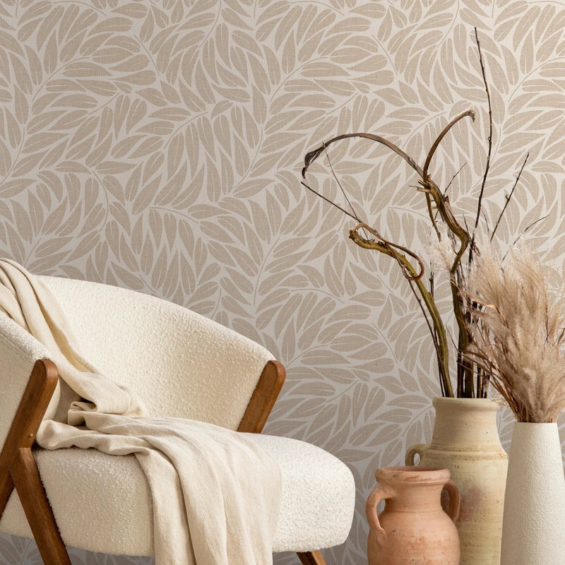 Marburg Boho 35796 Beige wallpaper with an overlapping leaf pattern shown behind a cream upholstered chair and ceramic vases holding dried branches and grasses.