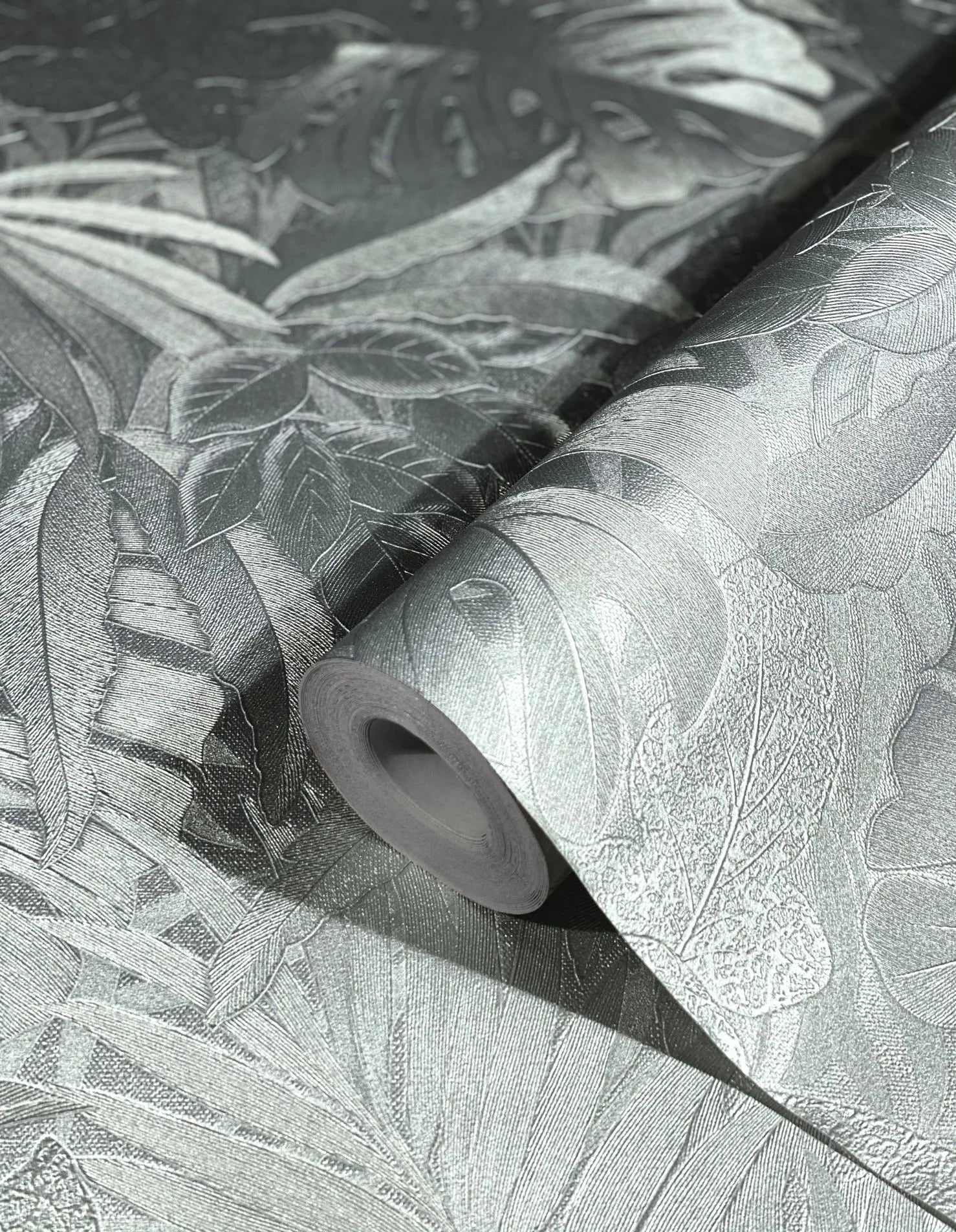 Marburg Botanica 33301 Close-up of a silver metallic botanical wallpaper roll on a matching unrolled sheet, showing embossed tropical leaf textures.