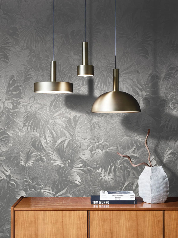 Marburg Botanica 33301 Silver-gray tropical leaf wallpaper in a styled room with three brushed brass pendant lights above a wooden credenza, plus stacked books and a white textured vase holding branches.