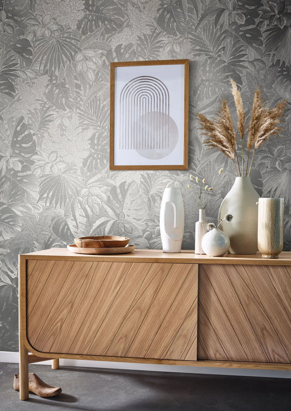 Marburg Botanica 33301 Silver botanical leaf wallpaper behind a wood sideboard styled with ceramic vases, pampas grass, wooden bowls, and a framed geometric print.