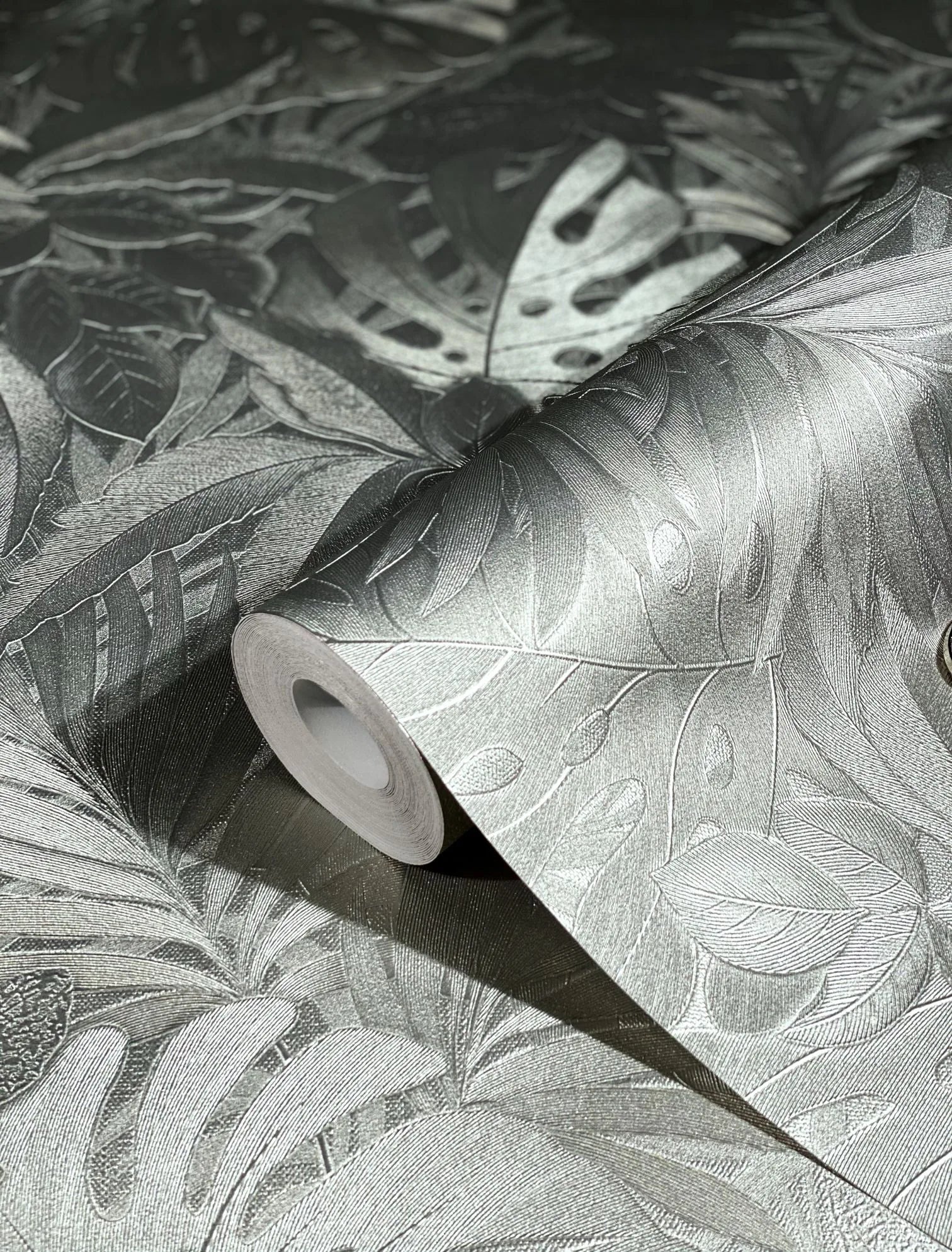 Marburg Botanica 33302 Close-up of a wallpaper roll showing an embossed tropical leaf pattern with a metallic sheen, resting on the same patterned surface.