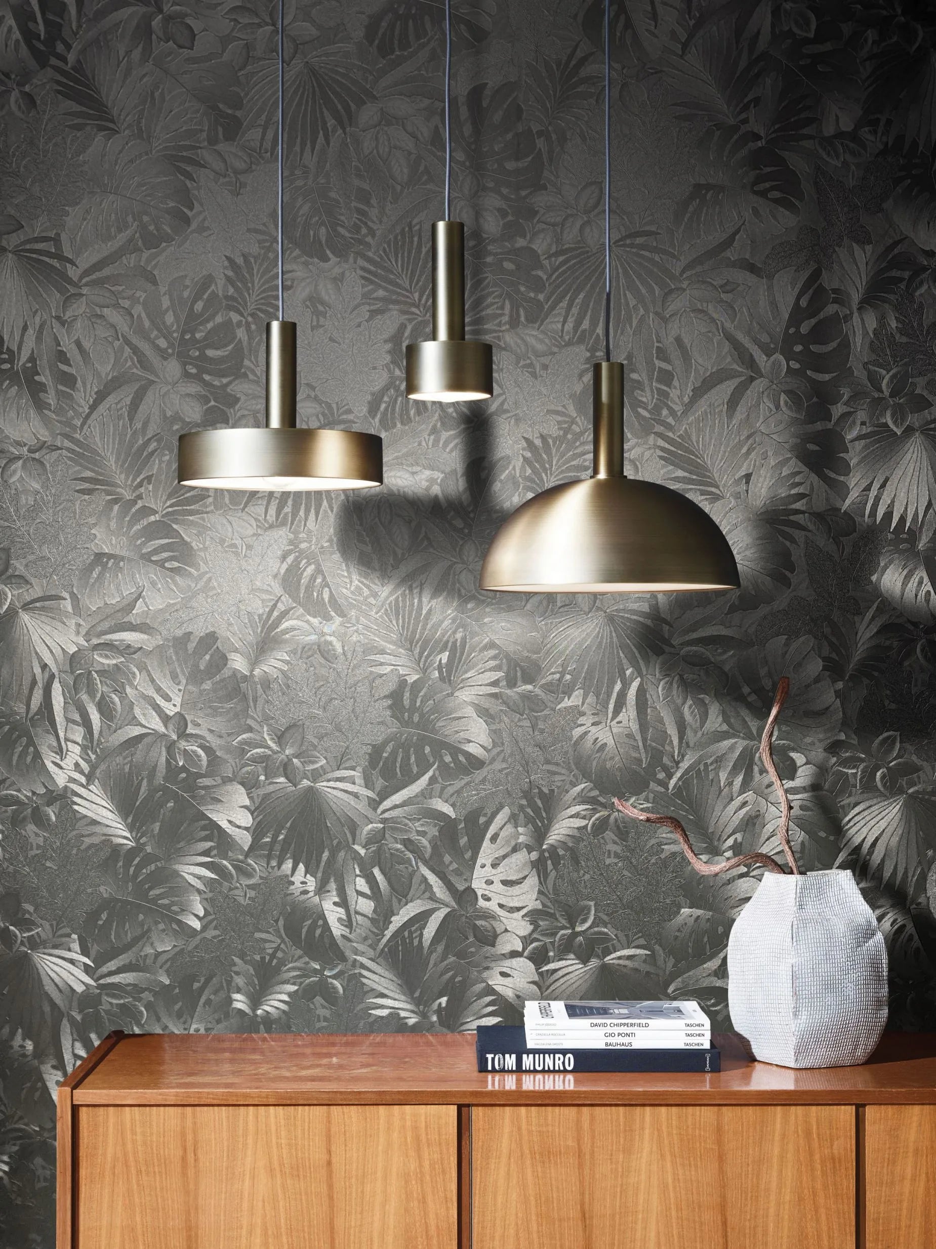 Marburg Botanica 33302 Tonal dark gray tropical leaf wallpaper shown in a styled scene with three metal pendant lights above a wood sideboard, stacked books, and a textured vase with branches.
