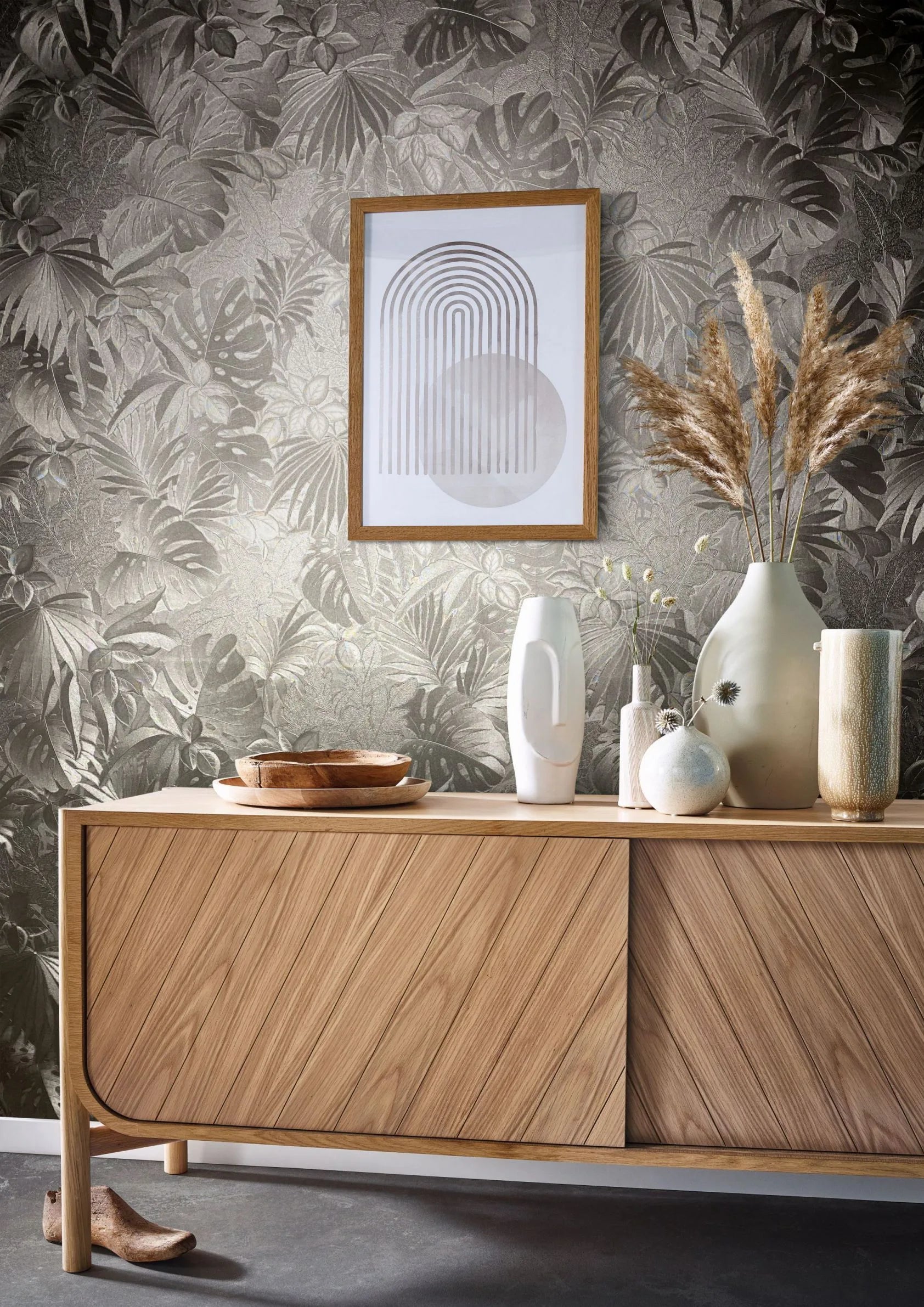 Marburg Botanica 33303 Tonal metallic botanical wallpaper with large tropical leaves shown behind a wooden sideboard topped with ceramic vases, pampas grass, wooden bowls, and a framed abstract print.
