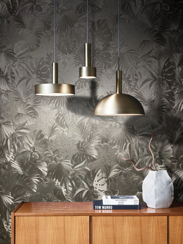 Marburg Botanica 33303 Tonal tropical leaf wallpaper with a subtle metallic sheen, shown behind three brass pendant lights, a wood cabinet, a small textured vase with branches, and a stack of books.