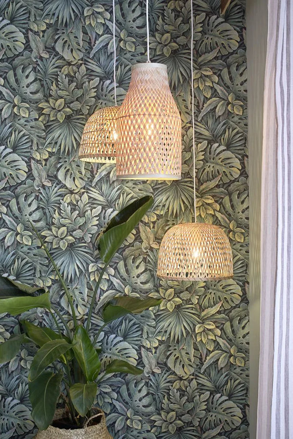 Marburg Botanica 33304 Tropical leaf wallpaper in green with layered monstera and palm motifs. Three woven rattan pendant lights hang in front, with a potted plant below and a striped curtain at the right edge.