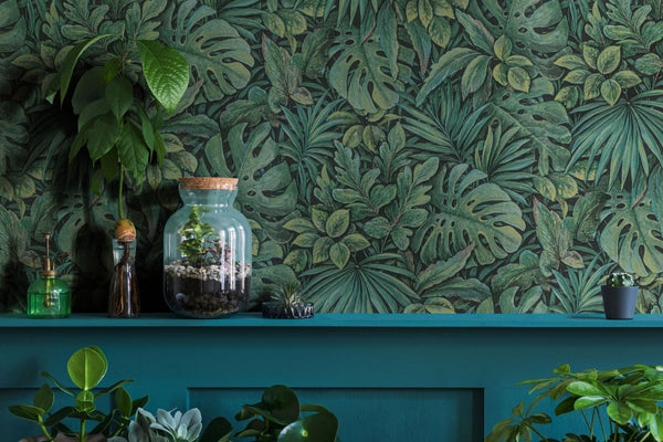 Marburg Botanica 33304 Green tropical leaf wallpaper with overlapping leaves behind a painted shelf styled with a glass terrarium jar, plant mister, bottle with rooted cutting, a small air plant, a tiny potted cactus, and other houseplants.