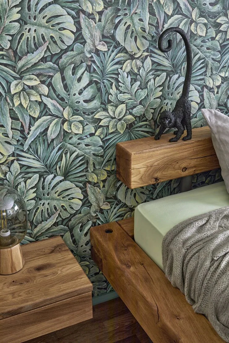Marburg Botanica 33304 Green tropical leaf wallpaper behind a wooden platform bed with a knit throw, a small monkey figurine on the headboard, and a glass-dome lamp on the nightstand.