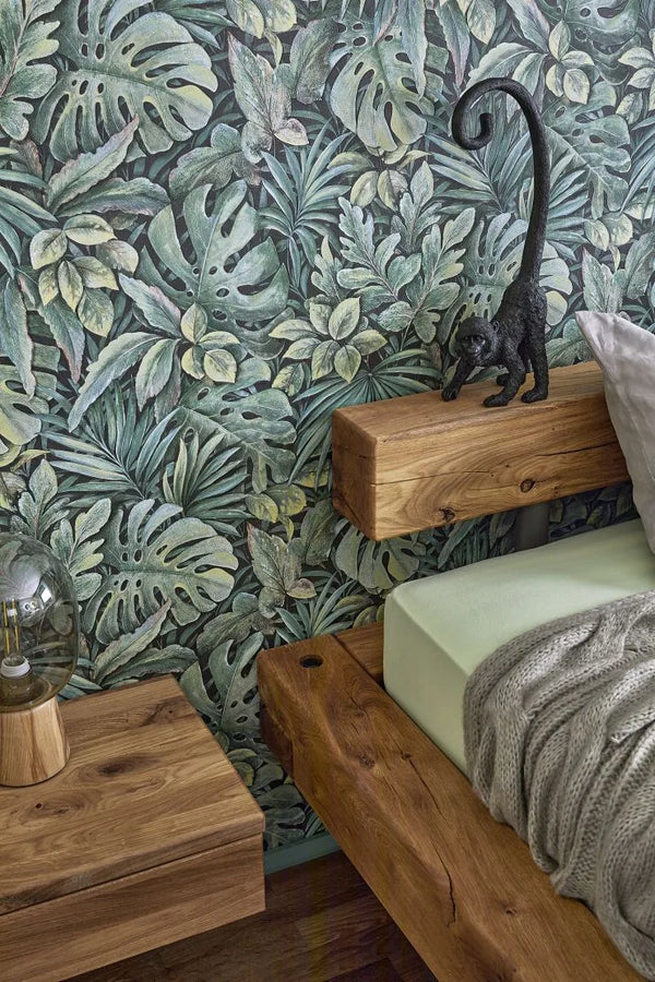 Marburg Botanica 33304 Green tropical leaf wallpaper behind a wooden platform bed with a knit throw, a small monkey figurine on the headboard, and a glass-dome lamp on the nightstand.