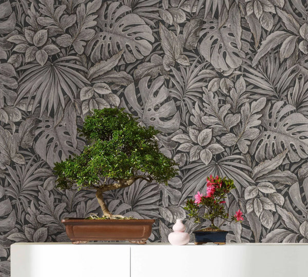 Marburg Botanica 33305 Dark monochrome tropical leaf wallpaper behind a white cabinet with a bonsai tree, a small pink vase, and a smaller flowering bonsai.