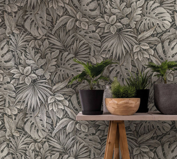 Marburg Botanica 33305 Tropical leaf wallpaper in muted neutral tones behind a narrow wooden console table holding several potted plants, a small clear bottle, a small green bottle, and a light wooden bowl.