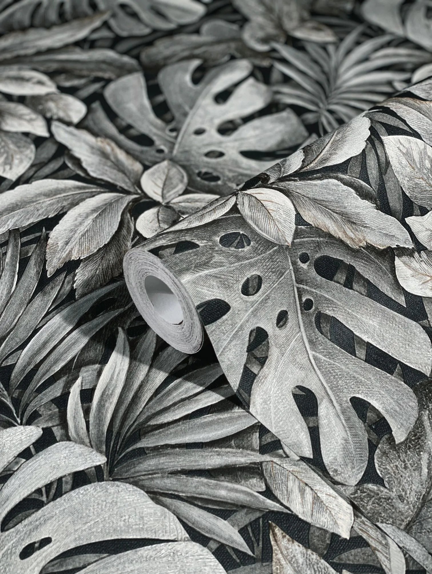 Marburg Botanica 33305 A wallpaper roll resting on the same pattern, showing large overlapping tropical leaves in gray tones on a dark background.