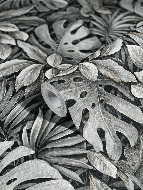 Marburg Botanica 33305 A wallpaper roll resting on the same pattern, showing large overlapping tropical leaves in gray tones on a dark background.
