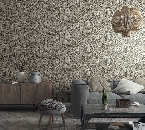 Marburg Botanica 33305 Wide living room scene featuring a neutral brown tropical leaf wallpaper with overlapping monstera and palm leaves behind a gray sofa, coffee table, sideboard, woven pendant light, and small decor.