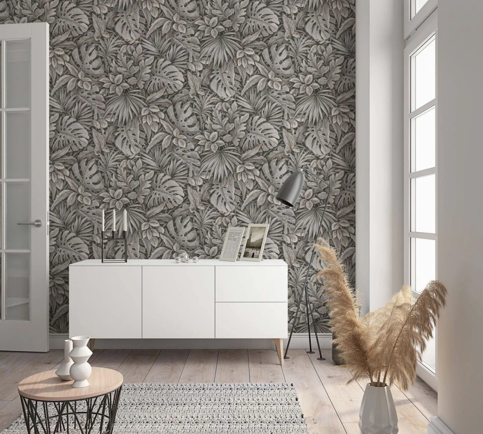 Marburg Botanica 33305 Room scene featuring a dense tropical leaf wallpaper in muted brown‑gray tones behind a white sideboard with decor, an angled floor lamp, a small round coffee table with a sculptural vase, a woven rug, and a vase of pampas grass beside a tall window.